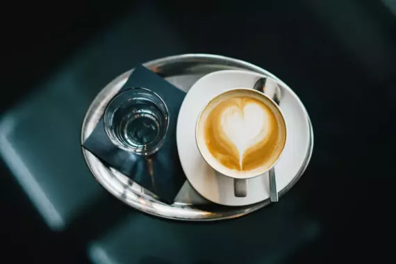 Ein frisch zubereiteter Cappuccino mit Herz-Latte-Art auf einem metallischen Tablett, serviert mit einem Glas Wasser und einer dunkelblauen Serviette. Die Aufnahme ist aus der Vogelperspektive aufgenommen und zeigt den Kontrast zwischen dem cremigen Schaum des Kaffees und der dunklen Oberfläche des Tisches, was eine einladende Pause für Kaffeeliebhaber suggeriert.