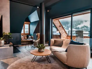 Ein heller Wohnbereich mit einem grauen Sofa und einem kleinen runden Holztisch, die auf eine, grauen Teppich stehen. Im Hintergrund ist eine weitere Sitzecke, ein Fernseher sowie eine Dachterrasse zu sehen.