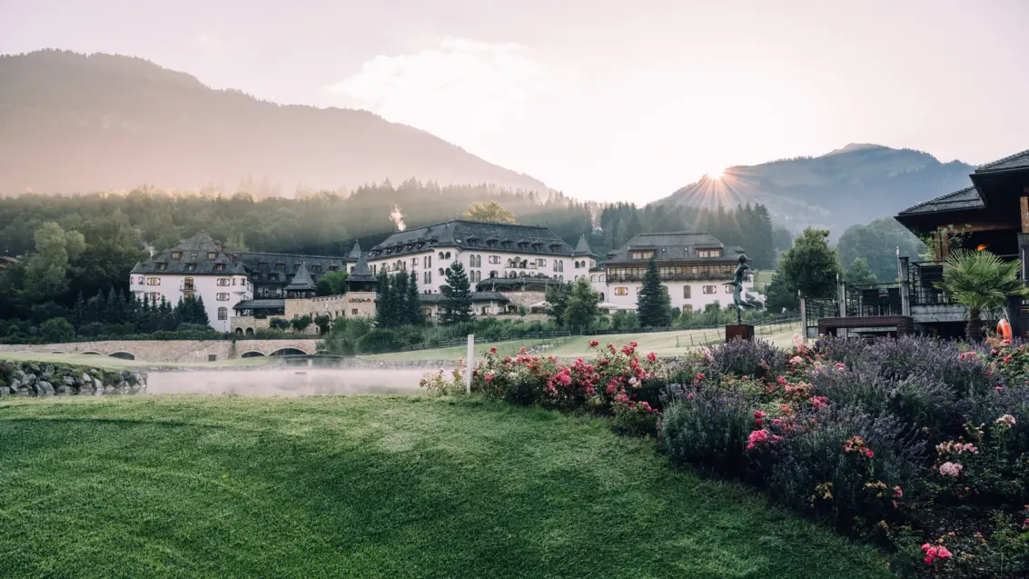Idyllische Ansicht des A-ROSA Hotels in Kitzbühel während eines nebligen Sonnenaufgangs mit den ersten Strahlen, die über die Bergspitzen streichen. Im Vordergrund ein gepflegter Rasen bepflanzt mit blühenden Rosen und Lavendel, der sich vor dem traditionellen alpinen Hotelgebäude erstreckt. Die ruhige Wasserfläche eines Sees reflektiert das sanfte Morgenlicht und betont die ruhige und erholsame Atmosphäre der Szenerie.