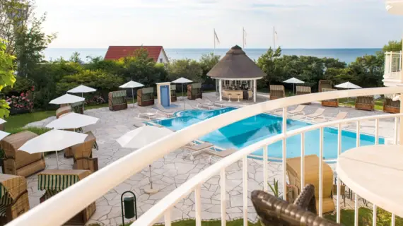 Blick von einem Balkon auf einen Pool mit Liegestühlen und Sonnenschirmen am Strand.