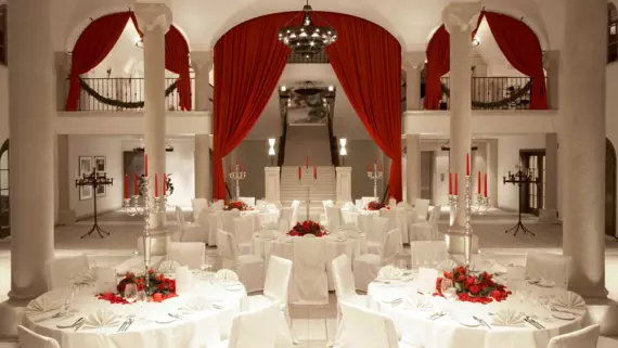 Eine große Halle mit Säulen, roten Vorhängen und vielen weißen Tischen.