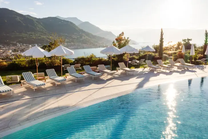 Pool mit Stühlen und Sonnenschirmen am Wasser, umgeben von Bergen und Bäumen.