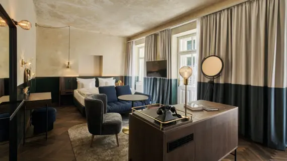 Stilvoll eingerichtetes Zimmer im Straubinger Grand Hotel Bad Gastein mit Designmöbeln, Doppelbett, Lounge-Bereich und Tageslicht durch große Fenster mit Vorhängen.