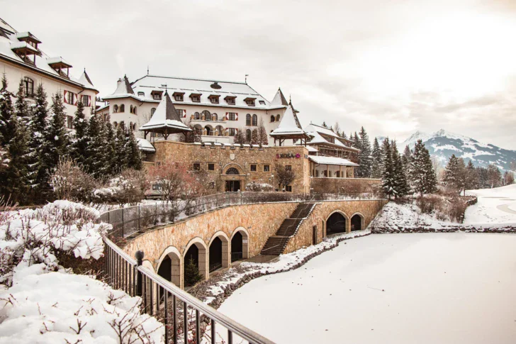 Ein Gebäude im Stil eines Schlosses mit einer Brücke über einem Teich in einer verschneiten Winterlandschaft.