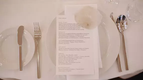 Ein elegant gedeckter Esstisch für eine Hochzeitsfeier mit einem weißen Teller, Besteck und einem bedruckten Menü. Das Menü ist kunstvoll auf dem Teller platziert, darüber liegt eine transparente Papierrolle mit dem Namen "Jan". Die Tischdekoration ist schlicht und stilvoll, perfekt für ein festliches Essen.