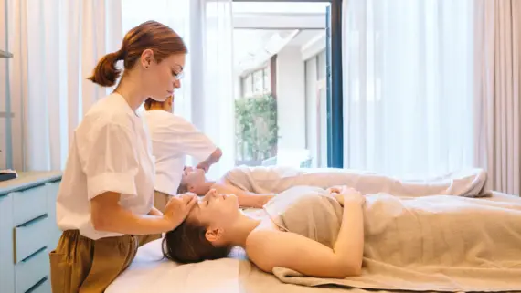 Two women lie relaxed on their backs while they each receive a facial treatment.