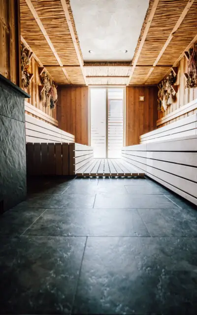 Eine Sauna aus Holz, die einladend und ruhig wirkt, mit mehrstufigen Sitzbänken auf beiden Seiten des Raumes. Die Wand ist mit natürlichen Hölzern verkleidet und mit getrockneten Kräuterbündeln geschmückt, die eine traditionelle und naturverbundene Atmosphäre schaffen. Der Boden ist mit dunklen Schieferfliesen ausgelegt, und eine Glassaunatür am Ende des Raumes lässt Tageslicht hereinströmen.