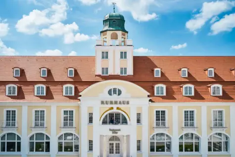 Außenansicht des A-ROSA Kurhaus Binz mit hellen Fassaden, einem roten Dach und Turm. 