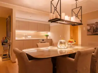 There is a large dining table with 6 upholstered chairs, a champagne cooler and a fruit bowl. In the background is a beige kitchen unit.