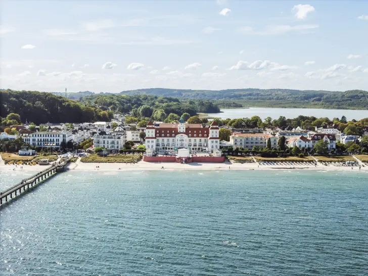 Großes weißes Gebäude an der Ostseeküste mit dem Meer im Vordergrund und mit Bäumen und einer Stadtkulisse im Hintergrund.