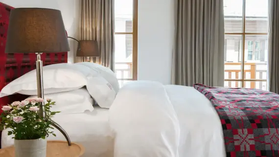 Ein rotes Polsterbett mit weißer Bettwäsche und einer roten Tagesdecke. Im Vordergrund ist ein Nachttisch mit einer Lampe und einer Blume zu sehen und im Hintergrund ein bodentiefes Fenster mit Zugang zu einem Balkon. 