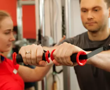 Ein Mann trainiert an einem Gerät, wobei er seine Arme mit einer Maschine vor seinem Körper ausgestreckt hält. Daneben steht eine Trainerin in rotem Oberteil. 