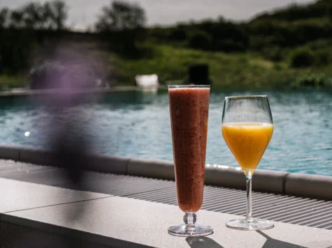 Zwei Gläser stehen auf einer Steinbank. Das eine Getränk ist in einem hohen Glas und rot, das andere ein Orangensaft. Im Hintergrund befindet sich ein Außenpool und grüne Landschaft. 