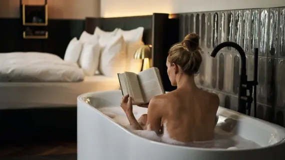 Frau liest ein Buch in einer Badewanne, im Hintergrund ein Hotelbett mit weißer Bettwäsche.