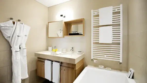 A bright bathroom with a bathtub and a vanity unit with two washbasins. There are two bathrobes hanging on hangers on the wall and a towel warmer above the bathtub.