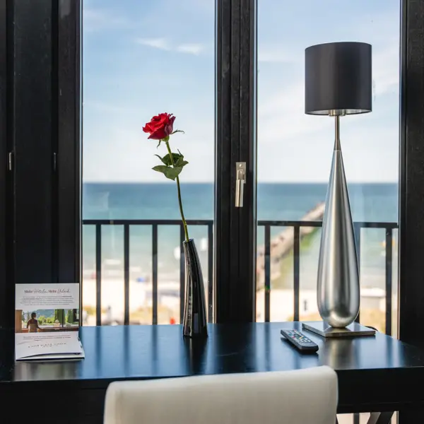 Ein dunkler Schreibtisch mit Blumenvase und silberner Lampe vor einem großen Fenster mit Blick auf die Ostsee. 