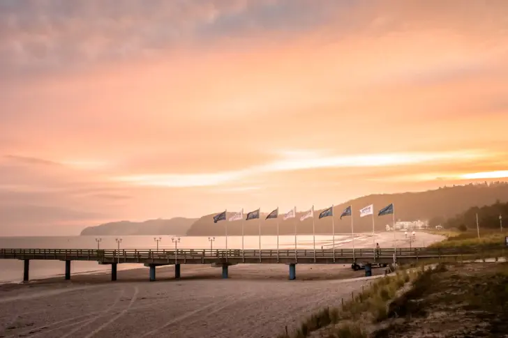 Über einen leeren Strand bei Sonnenaufgang führt ein langer Steg, auf dem Flaggen gehisst sind, auf das Wasser hinaus. 