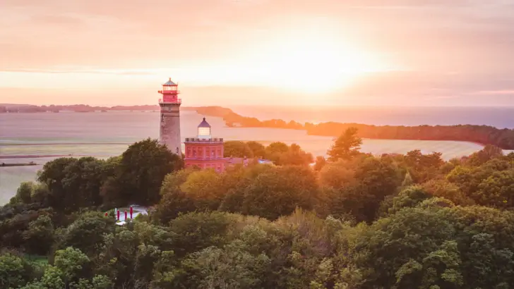 Leuchtturm inmitten eines Waldes bei Sonnenuntergang, umgeben von Bäumen und einem Gewässer.