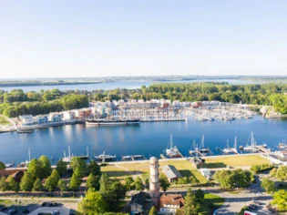 Ein Luftbild von Travemünde zeigt die Hafenanlage mit einer Vielzahl von Segelbooten und Schiffen, die im Yachthafen liegen. In der Mitte des Bildes ragt ein alter Leuchtturm hervor, umgeben von grünen Bäumen und Parkflächen. I