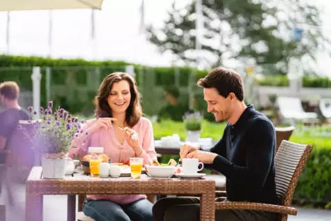 Ein Paar sitzt fröhlich an einem gedeckten Frühstückstisch draußen auf einer Terrasse. 