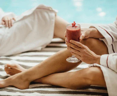 Zwei Personen sitzen auf einer gestreiften Oberfläche in Bademänteln, wobei die rechts sitzende Person ein Glas mit einem roten Getränk in der Hand hält. Im Hintergrund ist das Blau des Pools zu erkennen.   