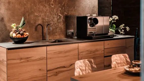 Eine Küchenzeile in Holzoptik mit einem Waschbecken, einer Kaffeemaschine und einem Obstkorb. Im Vordergrund ist ein Holztisch mit zwei Stühlen zu sehen.