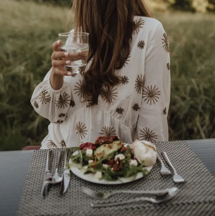 Eine Frau in einem weißen Blumenkleid sitzt an einem Tisch. Sie hält ein Glas Wasser in ihrer Hand, vor ihr steht ein Salatteller. 
