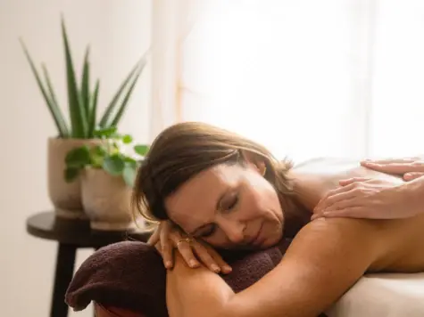 The picture shows a relaxed woman enjoying a soothing back massage. She is lying on her stomach with her head resting comfortably on her hands on a brown cushion. In the background you can see a peaceful ambience, characterized by soft light and two green potted plants on a stool made of dark wood, which contribute to the calming atmosphere. The massager's hands are gently placed on the person's shoulder blades.