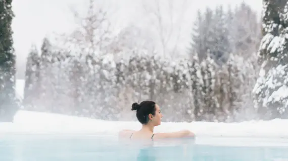 Eine Frau schwimmt in einem warm dampfenden Außenpool und blickt in die schneebedeckte Ferne. 