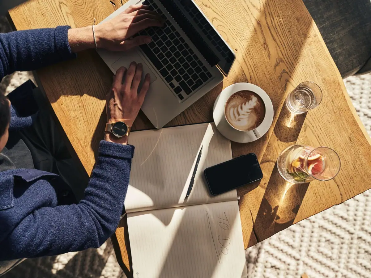 Eine Person benutzt einen Laptop auf einem Tisch und neben ihm liegt ein großes Notizbuch, ein Kaffee und ein Handy.
