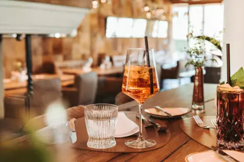  Ein elegant gedeckter Tisch in einem Restaurant mit einem Glas Roséwein, einem Wasserglas und einem Cocktail, umgeben von einer warmen, einladenden Atmosphäre und sanftem Tageslicht.