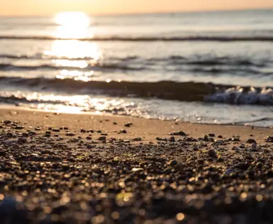 Eine Nahaufnahme eines Strandstückes, an dem die leichten Wellen des Wassers ruhig auf den Strand und kleine Steine trifft. Die tierstehende Sonne wird bald untergehen und spiegelt sich in warmen Farben auf dem Wasser.