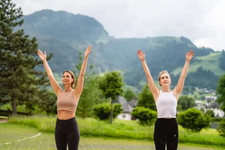 Zwei Frauen mit erhobenen Armen draußen, lächelnd in einer Yoga- oder Tanzpose.