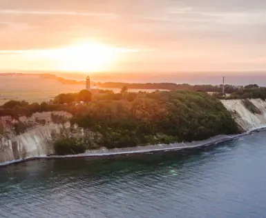 Ein Leuchtturm auf einer bewaldeten Klippe über der Ostsee bei Sonnenuntergang. Die Steilküste mit weißen Felsen trifft auf das ruhige, dunkelblaue Wasser, während die Sonne den Himmel in warme Farben taucht.