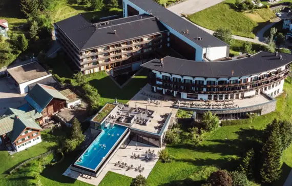 Large building surrounded by grassy areas and alpine-style houses, taken from a bird's eye view.