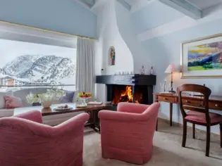 Ein Sofa und zwei Sessel stehen vor einem Kamin und durch ein Fenster ist die verschneite Bergwelt zu sehen.