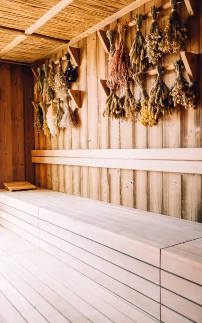  Das Bild zeigt das Innere einer Sauna mit natürlicher Holzverkleidung an Wänden und einer Reetverkleidung an der Decke. An der Wand hängen verschiedene Bündel aus getrockneten Kräutern, die zur Entspannung und Verbesserung des Raumduftes dienen könnten. Die Sauna ist mit langen, hellen Holzbänken ausgestattet, die zum Verweilen und Entspannen einladen. 