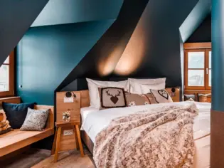 Ein gemütliches Bett mit einer braunen Kuscheldecke und drei Dekokissen im Alpenstil. Links neben dem Bett ist ein kleiner Nachttisch aus Holz und eine Sitzecke mit Kissen zu sehen.