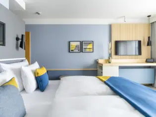 Ein Hotelzimmer mit einem großen Bett mit blau-gelber Tagesdecke und einem Schreibtisch im Hintergrund.