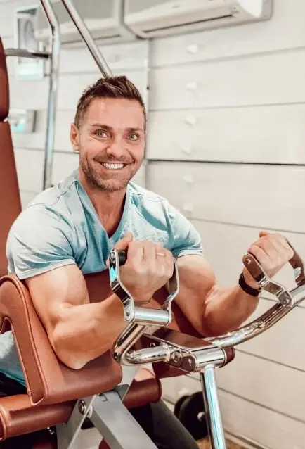 Ein Mann mit einem breiten Lächeln sitzt auf einem Fitness-Gerät und trainiert die Oberarme.