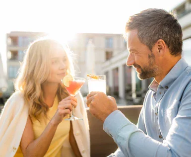 Ein Mann und eine Frau sitzen bei Sonnenschein draußen auf einer Terrasse und stoßen mit ihren Getränken lächelnd an. 