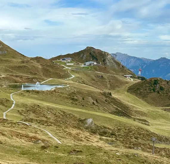 Ein Weg führt durch eine grüne Landschaft zu einem See, umgeben von Bergen und einem bewölkten Himmel.