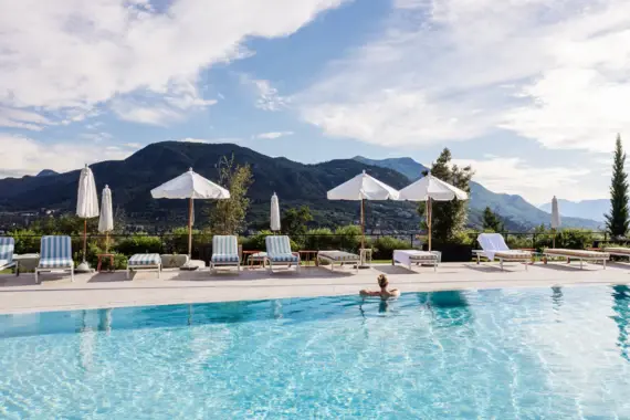 Eine Person lehnt am Beckenrand in einem Pool mit Sonnenschirmen, gestreiften Liegestühlen und Bergen im Hintergrund.