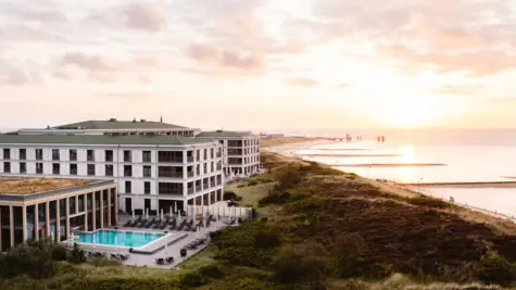 Ein Hotelgebäude mit einem Pool am Wasser, umgeben von Dünen und Strand.