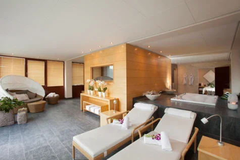 Eine private SPA-Suite mit hellen Liegen im Vordergrund und einem Whirlpool dahinter. Eine Holzsauna steht im Zentrum der Suite.