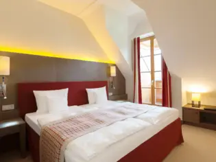 Ein Schlafzimmer mit einem roten Polsterbett, auf dem eine schmale karierte Tagesdecke liegt. Im Hintergrund ist ein bodentiefes Fenster zu sehen.