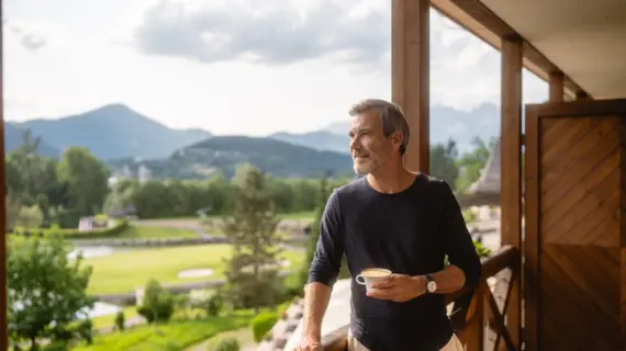 A man is standing on a balcony, holding a cup of coffee in his hand. He is looking into the distance at a mountain landscape.