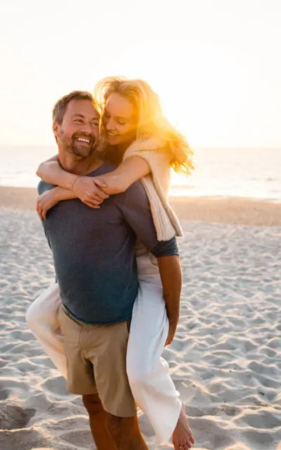 Mann trägt eine Frau auf seinem Rücken am Strand, beide lächeln.