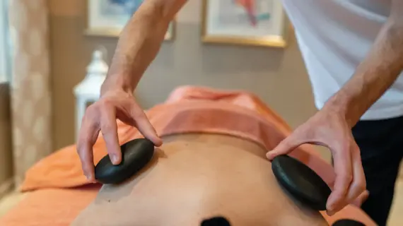 Eine Frau liegt auf einer Massageliege und ein Masseur hält zwei warme Steine auf ihren Rücken. 
