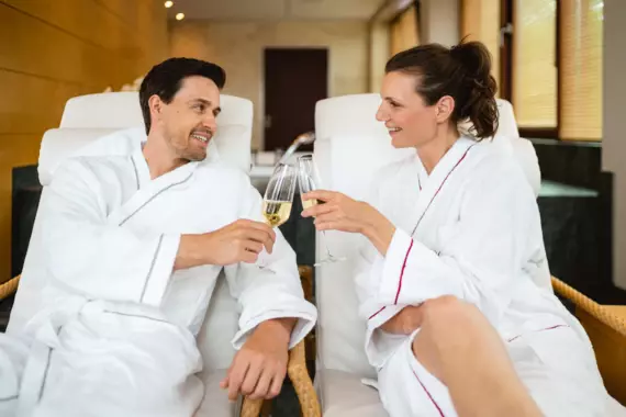 Zwei Personen in weißen Bademänteln, die entspannt sitzen und anstoßen, lächeln sich in einem wohligen Spa-Umfeld zu.